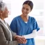 female caretaker in blue scrubs caring for and holding the hand of an elderly female patient