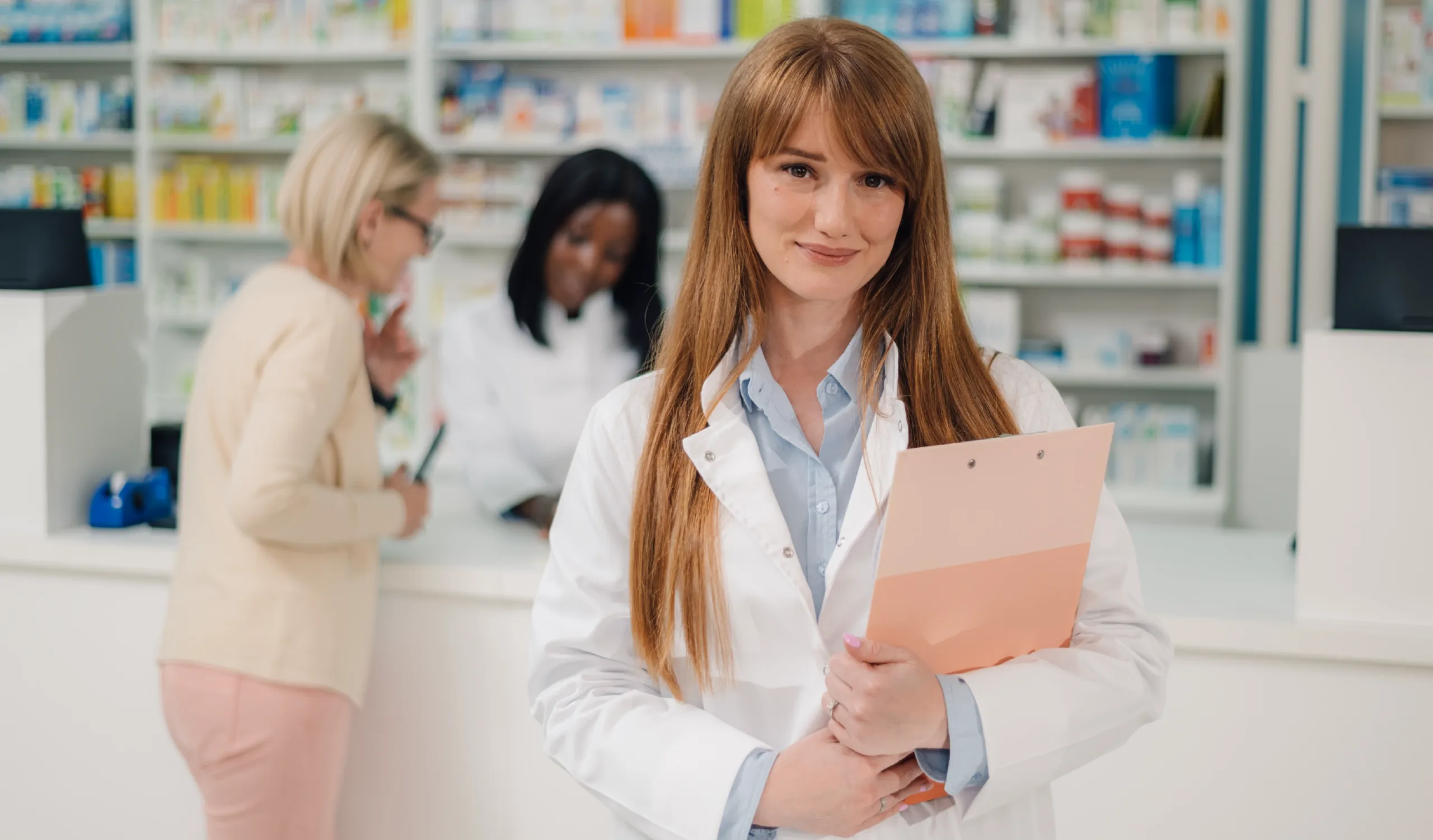 Pharmacy technician smiling politely while a coworker and client speak in the background of the pharmacy